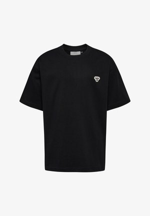 Lille barn står og smiler, iført en sort oversized t-shirt og bukser med blå sneakers, på en ensfarvet lys baggrund.