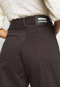 Jean en denim brun foncé à taille haute, avec des poches arrière et une étiquette à la taille. Tissu texturé avec des détails de couture visibles.