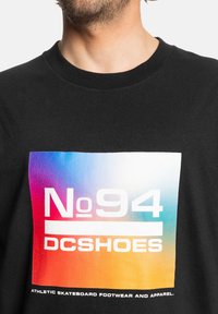 Zwarte katoenen t-shirt met een kleurrijke gradient graphic en vetgedrukte witte tekst "No 94" en "DC Shoes" gecentreerd op de voorkant.
