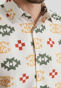 Chemise à manches courtes en tissu léger présentant un motif géométrique vert, rouge, jaune et orange sur fond blanc, avec un col pointu.