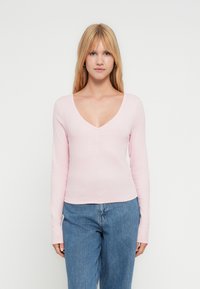 Νεαρή γυναίκα με μακριά ξανθά μαλλιά, που φοράει ροζ ανοιχτόχρωμο μπλουζάκι με V λαιμόκοψη και μακριά μανίκια και μπλε τζιν παντελόνι, μπροστά σε λευκό απλό φόντο.