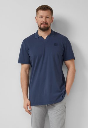 Mann mit Bart, der ein dunkelblaues kurzärmeliges Poloshirt und hellgraue Hosen trägt, steht vor einer schlichten hellgrauen Wand.