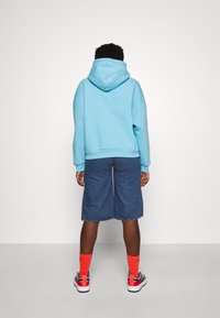 Sudadera azul claro con una capucha grande, combinada con pantalones cortos de mezclilla oscura. Calcetines naranjas brillantes y zapatillas multicolores completan el look.