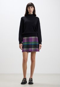 Maglione nero con collo alto; gonna a quadri viola e nera con righe verdi e blu; il modello indossa scarpe nere lucide.