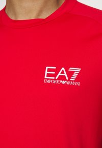 Rdeča kratka športna majica z dolgimi rokavi, izdelana iz zračnega materiala, s belim logotipom "EA7 EMPORIO ARMANI" spredaj. Teksturna površina.