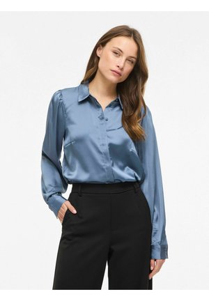 Kobieta ubrana w błyszczącą niebieską satynową bluzkę z długimi rękawami i czarne spodnie z wysokim stanem, pozująca z jedną ręką w kieszeni.