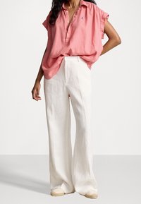 Camicia rosa in lino a maniche corte con colletto e bottoni, abbinata a pantaloni larghi in lino bianchi e scarpe espadrillas chiare.