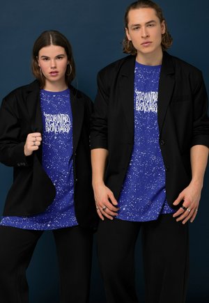 Dos modelos posan con trajes negros y camisas azules con estampado gráfico salpicado de blanco, sobre un fondo de estudio azul oscuro.
