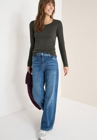 Langärmliges, dunkelgrünes Oberteil, kombiniert mit hellblauen, weiten Jeans. Auffällige silberne Hardware und Sneakers runden das legere Outfit ab.