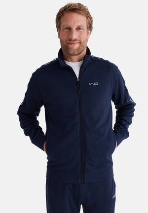 Lächelnder Mann mit Bart, der eine marineblaue Sportjacke mit Reißverschluss und passende Hose trägt und mit den Händen in den Taschen vor weißem Hintergrund steht.