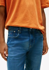 Persona que lleva pantalones de mezclilla azul desgastados y una camisa suelta de manga corta naranja, con las manos relajadas a los lados, cortada a la altura de la cintura.