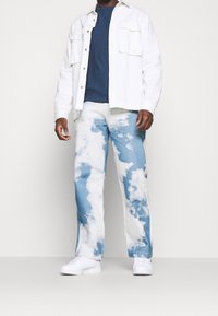 Vit långärmad skjorta över en marinblå t-shirt, i kombination med blå jeans med molnmönster och vita sneakers. Avslappnad passform och samtida design.