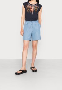 Laivasto sininen T-paita, jossa on värikkäitä höyhenpainatuksia, yhdistettynä vaaleansinisiin farkkushortseihin, joissa on joustava vyötärönauha ja sivutaskut. Mustat sandaalit.