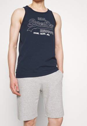 Młody mężczyzna w granatowej koszulce na ramiączkach marki SuperDry i jasnoszortach, stoi z jedną ręką w kieszeni.