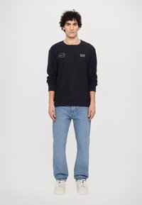 Jūras spēku sporta krekls ar izšūtiem logotipiem, apaļu kakla izgriezumu un ribotām piedurknēm, apvienots ar gaiši ziliem džinsiem un krēmkrāsas kedām. Vienkāršs fons.