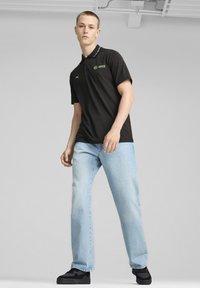 Sort polo med grønt logo, kortærmet og med krave, parret med lyseblå denim jeans og sorte sko.