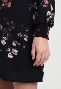Černé šaty s květinovými vzory v růžové a béžové barvě, s elastickými balónovými rukávy a texturovanými detaily na manžetách. Částečně viditelná ruka.