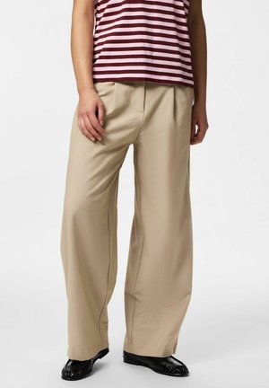 Person iført beige vidde bukser, sorte sko og en rød- og hvidstribet skjorte, stående mod en ensartet lys baggrund.