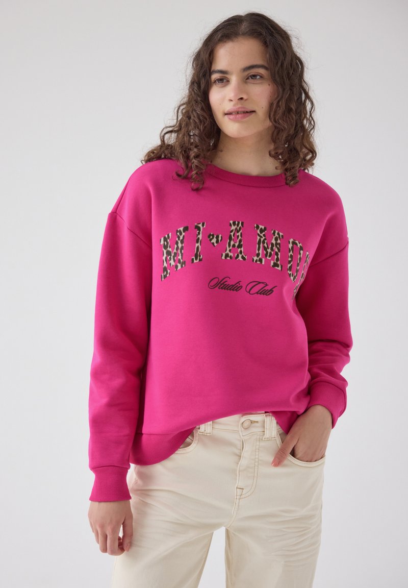 Femme aux cheveux bouclés portant un sweat-shirt rose vif avec un texte imprimé léopard "MI AMOR" et un pantalon couleur crème, main dans la poche.