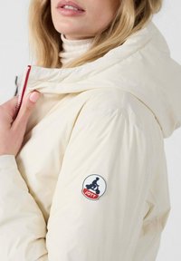 Chaqueta acolchada de color crema con un alto cuello y un parche de logo prominente en la manga. Tejido suave y brillante con detalles en cremallera roja.