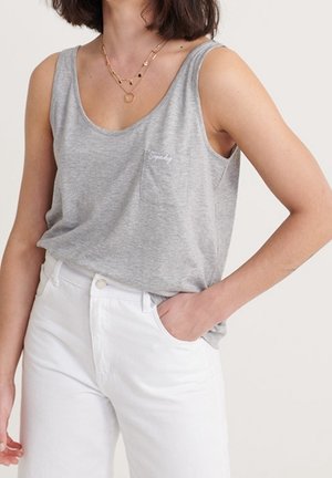 Woman wearing a grey sleeveless scoop neck top with a small chest pocket, paired with high-waisted white pants, hand in pocket.