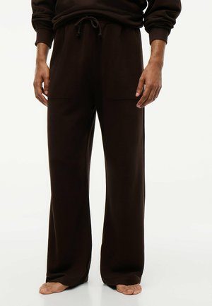 Man staat blootsvoets en draagt losse zwarte wijde broek met een koord in de taille en een bijpassend donker shirt met lange mouwen.