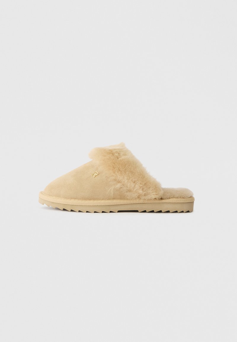 Beige mockasiner i mocka med mjuk pälsfoder, rund tå och struktur gummisula med zigzag-mönster. Guld logotypdetalj på sidan.