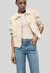Giacca beige cropped con colletto, chiusura frontale a bottoni, due tasche laterali e maniche arrotolate, abbinata a jeans a gamba dritta blu chiaro.