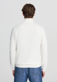 Pullover bianco in felpa con colletto alto e maniche lunghe, caratterizzato da un orlo a coste. Abbinato a jeans in denim blu.