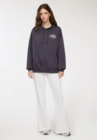 Felpa grigia scura con tasca frontale e lacci, con una grafica "FEELIN' LUCKY". Abbinata a pantaloni bianchi a campana e sneakers argento.