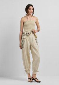 Femme portant un haut beige sans bretelles avec des volants et un pantalon ample assorti avec une broderie florale, debout devant un fond uni.