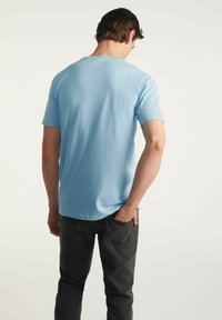 Jasnoniebieska bawełniana koszulka z okrągłym dekoltem i krótkimi rękawami, pokazana z tyłu, w zestawieniu z ciemnoszarymi spodniami. Gładka tekstura, prosty design.