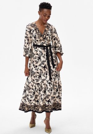 Kobieta ubrana w kremową sukienkę w czarne kwiatowe wzory, z rękawami o długości 3/4, czarnym paskiem w talii i złotymi szpiczastymi szpilkami, stojąca na białym tle.