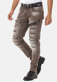 Ausgebleichte braune Jeans mit abgewetzten Patches und slim fit. Enthält ein schwarzes Logopatch und verzierte Beschläge am Bund. Getragen mit schwarzen Stiefeln.