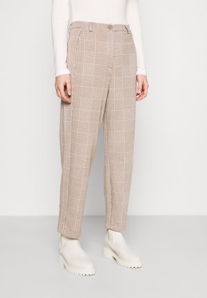 Personne portant un pantalon à carreaux beige et marron, un haut blanc à manches longues et des bottines blanches épaisses, debout sur un fond uni.