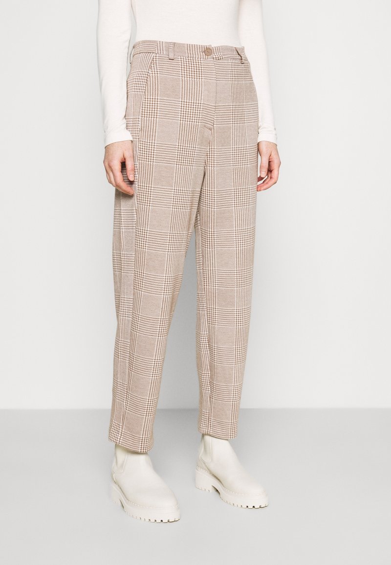 Personne portant un pantalon à carreaux beige et marron, un haut blanc à manches longues et des bottines blanches épaisses, debout sur un fond uni.