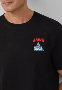 Zwart katoenen T-shirt met een geborduurd "JAWS" logo en een haai ontwerp in rood en blauw, met lichtblauwe waterspetters.