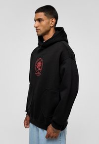 Zwarte hoodie met een voorzak, voorzien van een rode roos grafiek op de borst. Gemaakt van zachte stof met een relaxed fit en een capuchon met trekkoord.