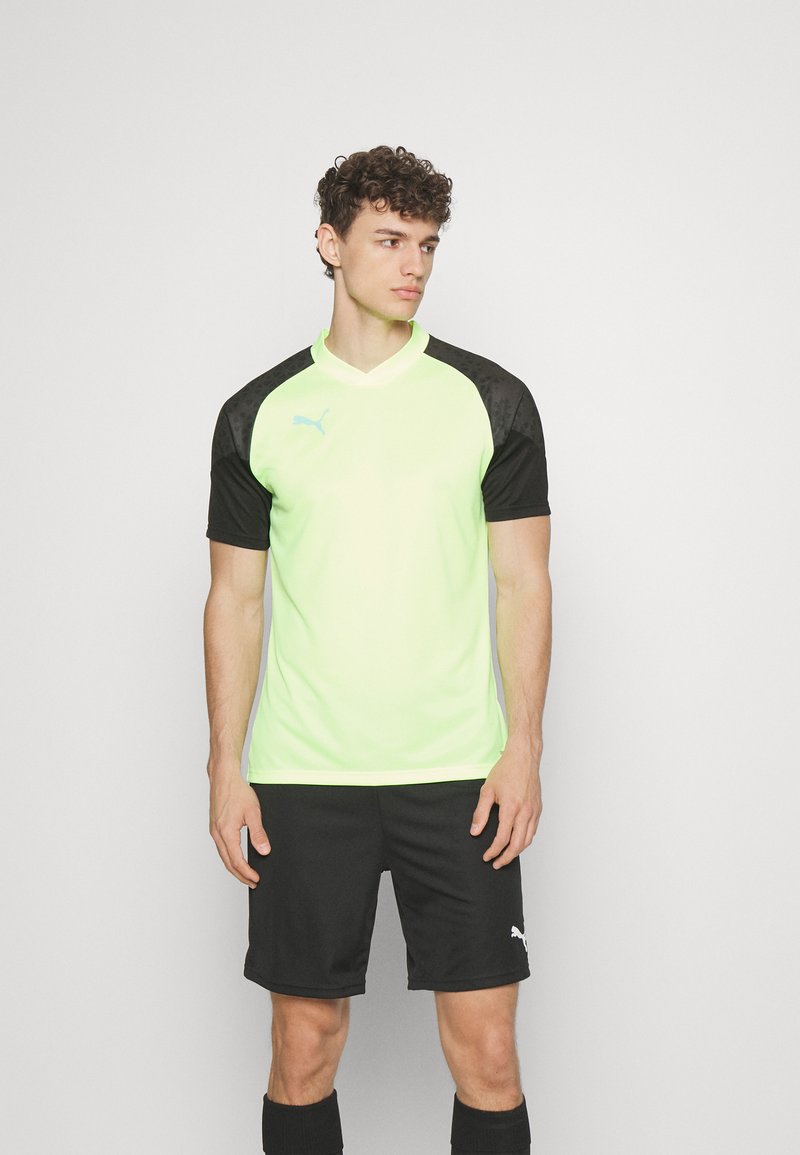 Camiseta de fútbol de manga corta en un brillante color verde neón, con mangas raglán negras y el logo de PUMA en blanco. Combinada con pantalones cortos y calcetines negros.