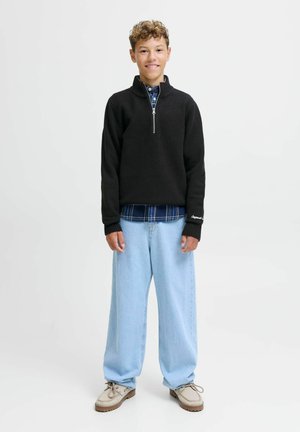Pullover in pile nero con colletto zip, indossato sopra una camicia a quadri blu, abbinato a jeans di jeans a gamba larga blu chiaro e scarpe beige.