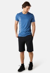 Blå atletisk t-shirt med logotyp på sidan, ihop med svarta shorts och svarta sneakers. Tyget verkar vara mjukt och lätt.