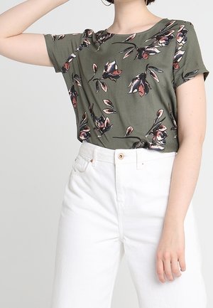 Persoon draagt een olijfgroen kort mouwtopje met roze en zwarte bloemenprints, gecombineerd met witte broek met hoge taille.
