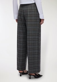 Donna che indossa pantaloni larghi a quadri grigi, una camicia bianca a maniche lunghe e scarpe con tacco a slittino nere, vista di spalle.