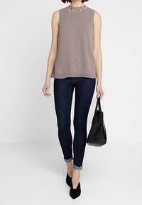 Blouse sans manches couleur mauve doux avec un col montant et des perles décoratives. Associée à un jean skinny bleu foncé et un sac à main noir texturé.