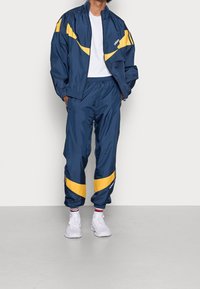 Marinblå vindjacka med gula detaljer, parat med matchande byxor. Tillverkad av glänsande, lätt material, har elastiska muddar och midjeband.