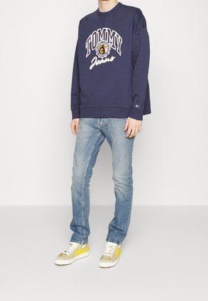 Sudadera azul marino con letras blancas "TOMMY Jeans", combinada con vaqueros azul claro y zapatillas beige con detalles amarillos. Corte relajado, estilo casual.