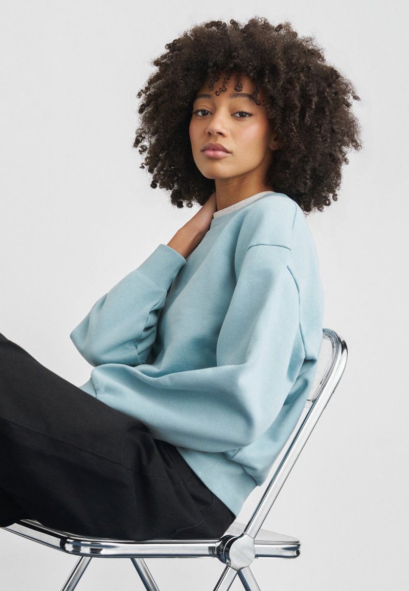 Ανοιχτό μπλε φούτερ με χαλαρούς ώμους και ριμπ λαιμόκοψη, σε συνδυασμό με μαύρο παντελόνι, καθισμένος σε καρέκλα από χρώμιο μπροστά σε λευκό φόντο.