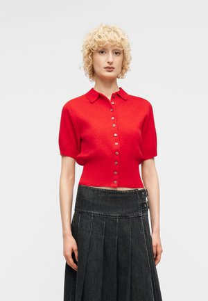 Ξανθό άτομο που φοράει κόκκινο κοντομάνικο πουκάμισο με κουμπιά και ψηλόμεση μαύρη πλισέ φούστα με αγκράφες, στέκεται μπροστά σε λευκό φόντο.