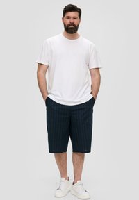 T-shirt blanc à manches courtes, shorts navy à rayures, et baskets blanches. Coupe décontractée avec un design relax. Fond uni.