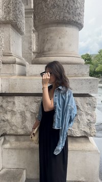 Τζιν μπουφάν πάνω από ένα μαύρο μακρύ φόρεμα, κρατώντας μια ελαφριά τσάντα. Το φόντο περιλαμβάνει υφασμάτινους πέτρινους κίονες και πράσινη βλάστηση.